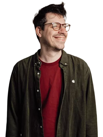 Een man met kort bruin haar en een bril lacht naar de camera. Hij draagt een roze-rood t-shirt en staat tegen een effen witte achtergrond.