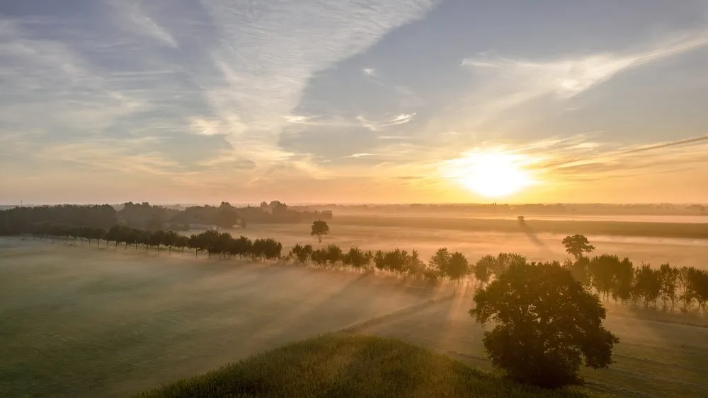 Zonsopgang boven een mistig landelijk landschap met bomen en velden onder een gedeeltelijk bewolkte hemel.