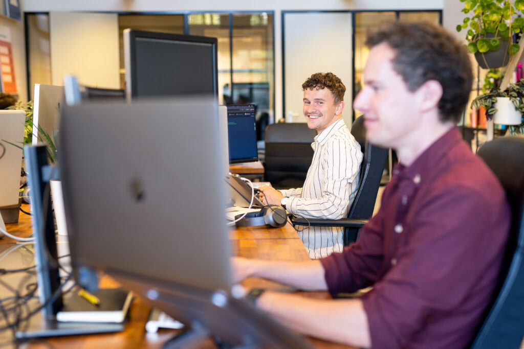 Nino en Jeroen werkend op het kantoor achter hun bureau. Nino lacht de camera in