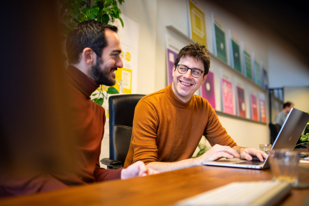 Aron is scherp in beeld terwijl hij lachend werkt achter zijn laptop. Werner zit ernaast en kijkt naar Aron, beiden in een ontspannen kantooromgeving.