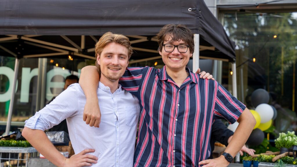 Frank en Max staan lachend naast elkaar buiten, met de armen om elkaars schouders.