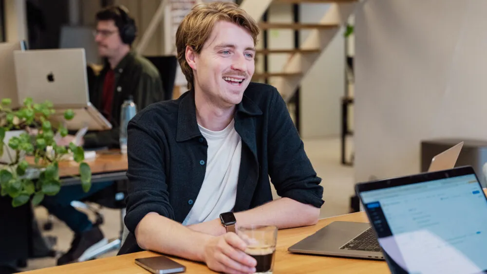 Lachende man met een glas in zijn hand aan een bureau met laptops in een modern kantoor.