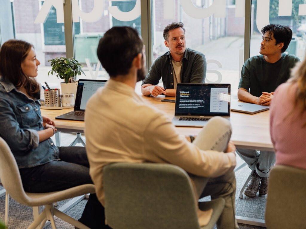 Mensen zitten met laptops rond een tafel en zijn in gesprek.
