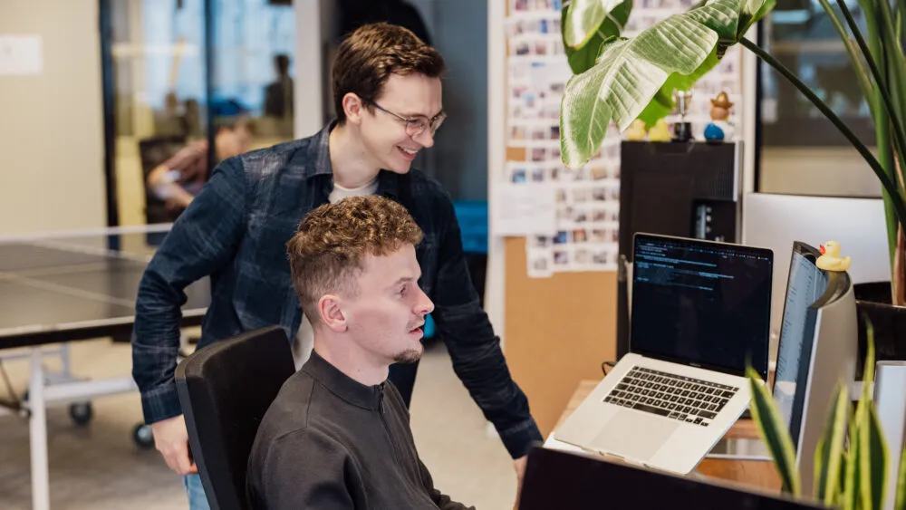 Twee mensen aan een bureau kijken naar code op een laptop, één zittend en één staand.