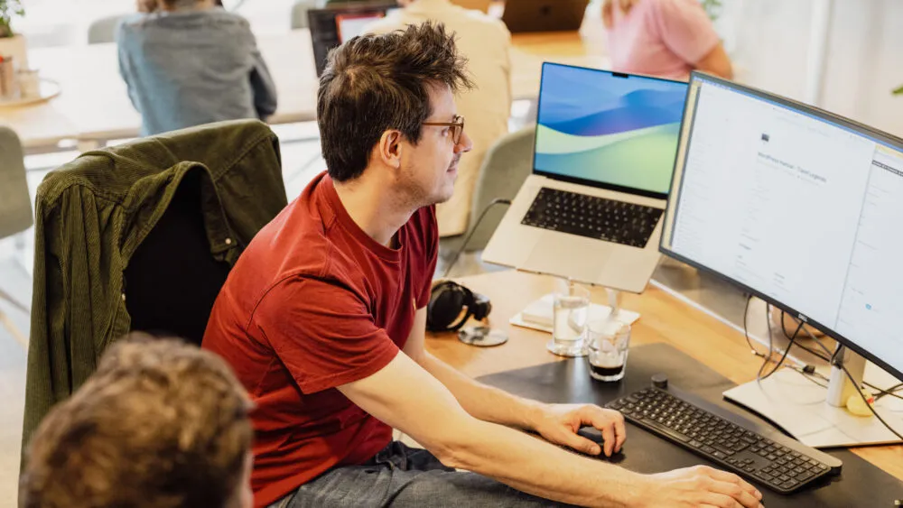 Man in rood shirt werkt aan een bureau met meerdere monitoren in een kantooromgeving.