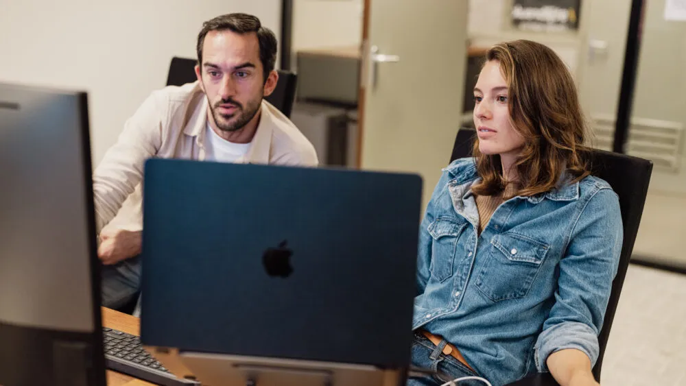 Twee mensen werken samen aan een bureau met computers in een kantooromgeving.