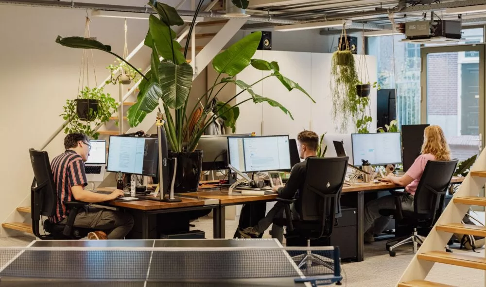 Een kantoor met vier mensen die aan een bureau werken, omringd door planten en een pingpongtafel.