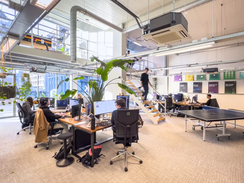 Modern kantoor met mensen die achter een bureau werken, een persoon op een trap en planten eromheen.
