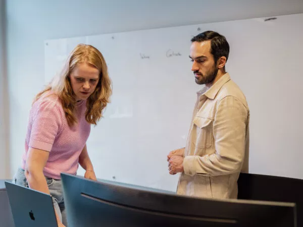 Twee mensen werken samen aan een computer op kantoor, met een whiteboard achter zich.