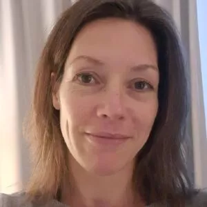 Vrouw met lichtbruin haar, licht glimlachend, staand binnen met een gordijn op de achtergrond.