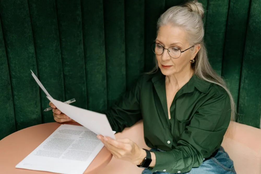 Oudere vrouw met bril leest onderzoeksdocumenten aan een tafel, draagt een groen shirt.