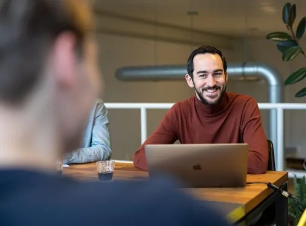 Man in een rode trui glimlacht tijdens een vergadering, zittend aan een tafel met een laptop.