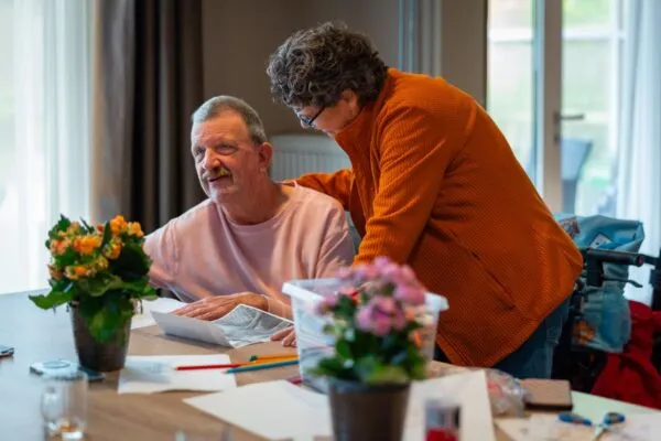 Oudere vrouw biedt ondersteuning aan een man aan een tafel met bloemen en kunstbenodigdheden.