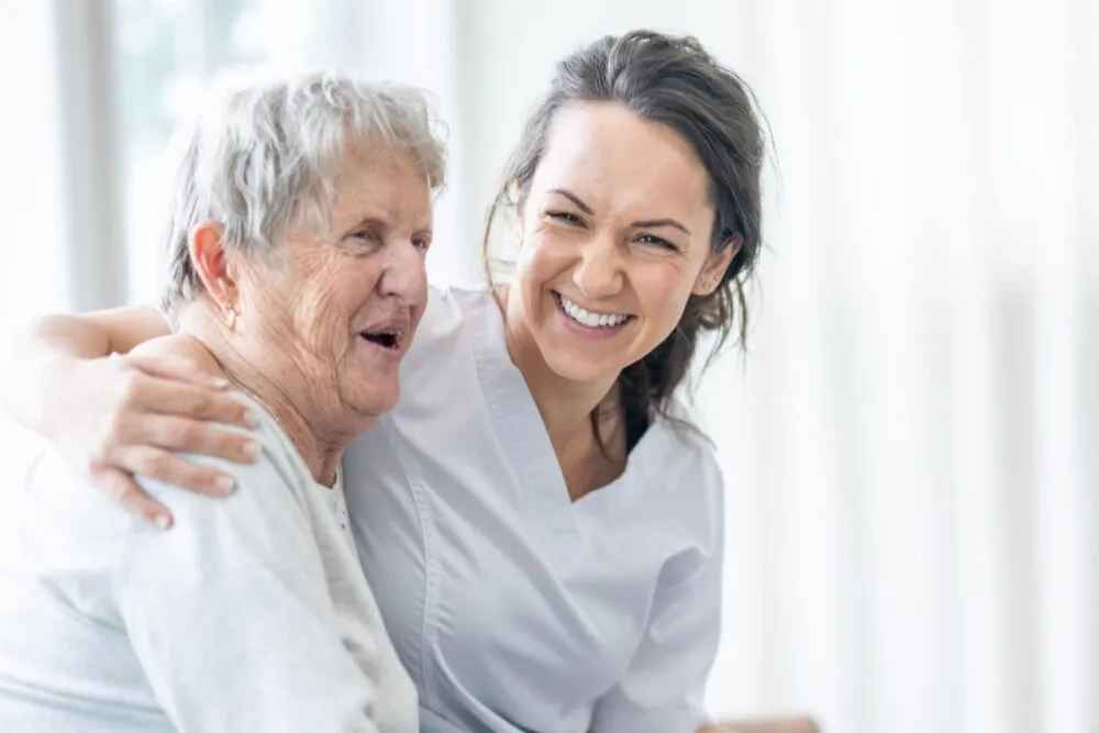 Glimlachende verpleegster die een oudere vrouw omhelst, beiden blij en vrolijk met een dempact touch.